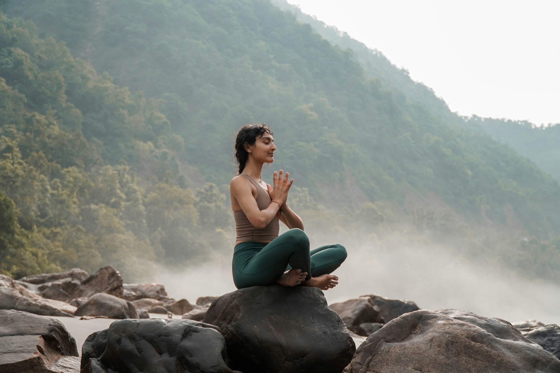 A person sitting in calm meditation on river rocks among misty green mountains